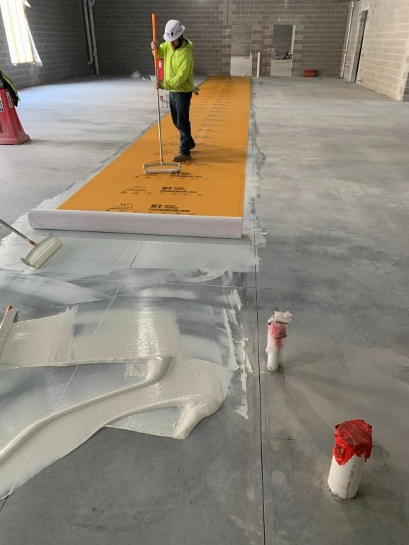 Construction worker in a hard hat and high-visibility shirt applies adhesive and rolls out bright orange temporary floor protection along a concrete floor inside an unfinished building, with exposed block walls and plumbing pipes visible in the foreground.
