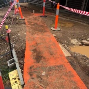 Muddy construction site walkway with orange ground protection mat surrounded by cones and caution tape, showing heavy dirt and foot traffic