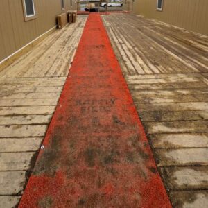 A long orange mat stretches across a weathered wooden walkway, flanked by brown walls. 