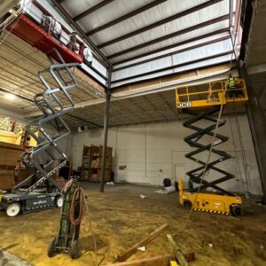 Two scissor lifts in a warehouse work on overhead beams. The red and yellow lifts extend upwards, with workers repairing. Equipment and shelves in the background.