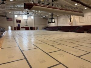 An empty school gymnasium with taped floor markings, basketball hoops, maroon bleachers, and U.S. flags; bright overhead lights give the space a quiet, spacious feel.