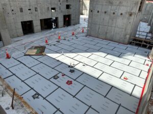 A concrete building interior has a floor covered in labeled protective panels, with orange safety cones, tools, and workers nearby.